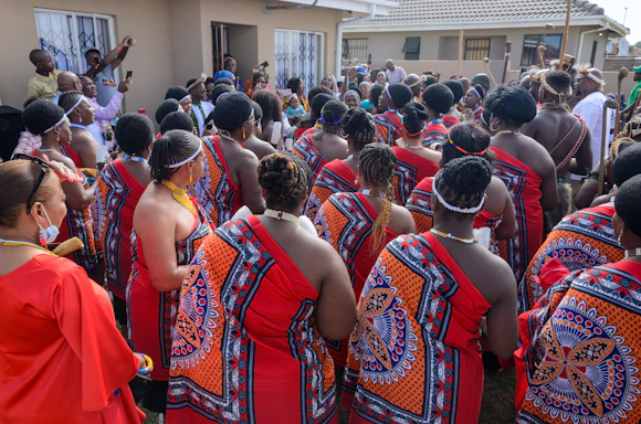 A group of people gathered outside a building, wearing traditional attire with bright red and intricate patterns. Many have braided hairstyles and some are adorned with beaded jewelry. The setting appears to be a cultural or social event.
