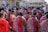 A group of people gathered outside a building, wearing traditional attire with bright red and intricate patterns. Many have braided hairstyles and some are adorned with beaded jewelry. The setting appears to be a cultural or social event.