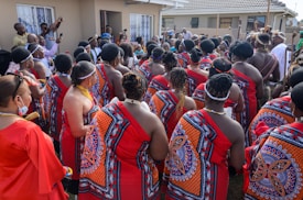 A group of people gathered outside a building, wearing traditional attire with bright red and intricate patterns. Many have braided hairstyles and some are adorned with beaded jewelry. The setting appears to be a cultural or social event.
