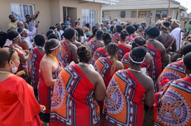 A group of people gathered outside a building, wearing traditional attire with bright red and intricate patterns. Many have braided hairstyles and some are adorned with beaded jewelry. The setting appears to be a cultural or social event.