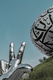 A metallic sculpture of a hand with three raised fingers is juxtaposed against a futuristic, orb-like structure adorned with intricate calligraphy. The scene is set against a clear blue sky with a slight green-tinged landscape, adding a surreal quality to the composition.