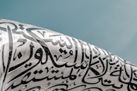 Arabic calligraphy intricately adorns the surface of a modern architectural structure with a sleek, metallic finish under a clear sky.
