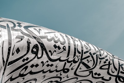 Arabic calligraphy intricately adorns the surface of a modern architectural structure with a sleek, metallic finish under a clear sky.