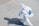 A person wearing a full protective suit, complete with a hood and respirator, is walking on a tiled surface. They are carrying a blue spray apparatus, likely used for disinfecting or sanitizing purposes. The shadow of the person is clearly visible on the ground.