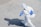 A person wearing a full protective suit, complete with a hood and respirator, is walking on a tiled surface. They are carrying a blue spray apparatus, likely used for disinfecting or sanitizing purposes. The shadow of the person is clearly visible on the ground.