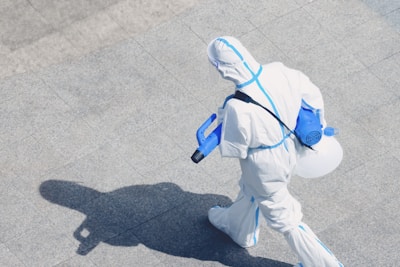 A person wearing a full protective suit, complete with a hood and respirator, is walking on a tiled surface. They are carrying a blue spray apparatus, likely used for disinfecting or sanitizing purposes. The shadow of the person is clearly visible on the ground.