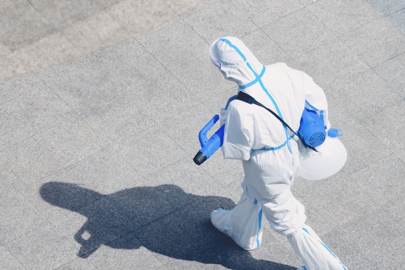 A person wearing a full protective suit, complete with a hood and respirator, is walking on a tiled surface. They are carrying a blue spray apparatus, likely used for disinfecting or sanitizing purposes. The shadow of the person is clearly visible on the ground.