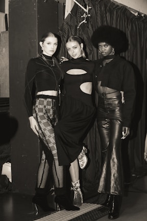 Three individuals are posing in fashionable attire, each wearing a unique style of black clothing. The setting appears to be indoors, with a curtain backdrop and ambient lighting. The mood is stylish and sophisticated.