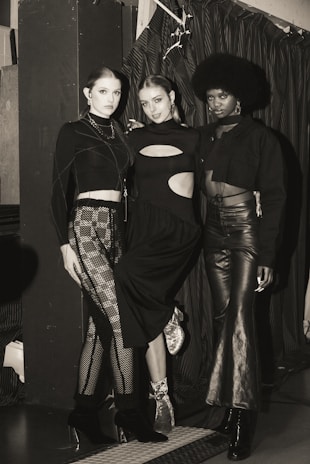 Three individuals are posing in fashionable attire, each wearing a unique style of black clothing. The setting appears to be indoors, with a curtain backdrop and ambient lighting. The mood is stylish and sophisticated.