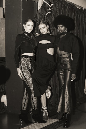 Three individuals are posing in fashionable attire, each wearing a unique style of black clothing. The setting appears to be indoors, with a curtain backdrop and ambient lighting. The mood is stylish and sophisticated.