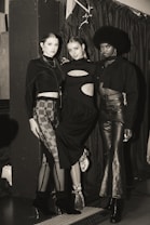 Three individuals are posing in fashionable attire, each wearing a unique style of black clothing. The setting appears to be indoors, with a curtain backdrop and ambient lighting. The mood is stylish and sophisticated.
