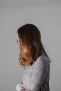 A person with long brown hair is seen in profile against a neutral gray background. The individual is wearing a checkered outfit and appears to have their arms crossed. The lighting is soft, creating a calm and introspective atmosphere.
