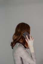 A person with long brown hair is holding a smartphone to their ear. They are wearing a plaid-patterned jacket and facing away from the camera, so their face is not visible. The background is plain and light gray, creating a minimalist setting.