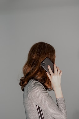 A person with long brown hair is holding a smartphone to their ear. They are wearing a plaid-patterned jacket and facing away from the camera, so their face is not visible. The background is plain and light gray, creating a minimalist setting.