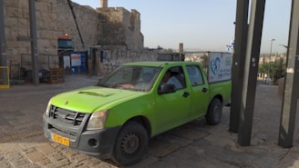 A bright green pickup truck is parked on a cobblestone path near an ancient stone wall. The truck has a yellow license plate and a sign on the side with blue graphics and text. Nearby, there is construction equipment and signs in front of the stone wall. The area is bathed in natural daylight.