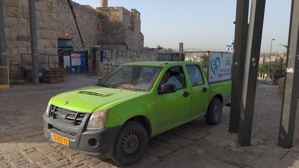 A bright green pickup truck is parked on a cobblestone path near an ancient stone wall. The truck has a yellow license plate and a sign on the side with blue graphics and text. Nearby, there is construction equipment and signs in front of the stone wall. The area is bathed in natural daylight.