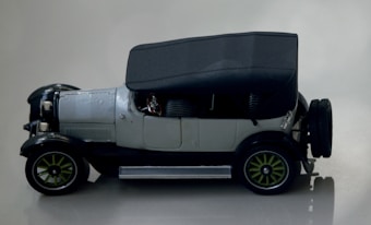 A vintage car model features a black canopy top, silver body, and green spoked wheels. The left side of the car is visible, showcasing intricate detailing on the hubcaps and hood. The setting is minimalist, with a neutral background that highlights the car's design.