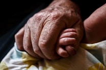 A close-up shot of an elderly hand gently holding a baby's hand. The image captures the contrast between the wrinkled, aged skin of the elder and the smooth, small hand of the baby, highlighting a moment of tenderness and connection. The background appears dark and out of focus, drawing attention to the two hands.