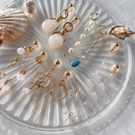 A carefully arranged collection of various elegant earrings set on a textured, round translucent platter. The earrings feature elements such as gold chains, pearls, turquoise stones, and crystal-like embellishments, accompanied by seashells as decorative accents.