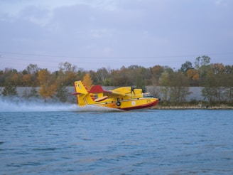 A yellow and red amphibious aircraft is skimming over a body of water with trees in the background. The aircraft is touching the water, creating a splash as it moves at high speed.