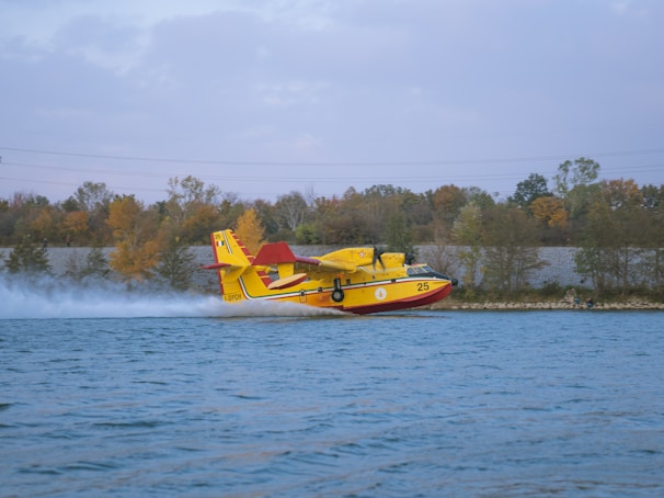 A yellow and red amphibious aircraft is skimming over a body of water with trees in the background. The aircraft is touching the water, creating a splash as it moves at high speed.
