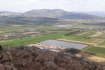 A vast rural landscape featuring agricultural fields with varying shades of green and brown, interspersed with small bodies of water. Rocky foreground with hills in the background under a partly cloudy sky.