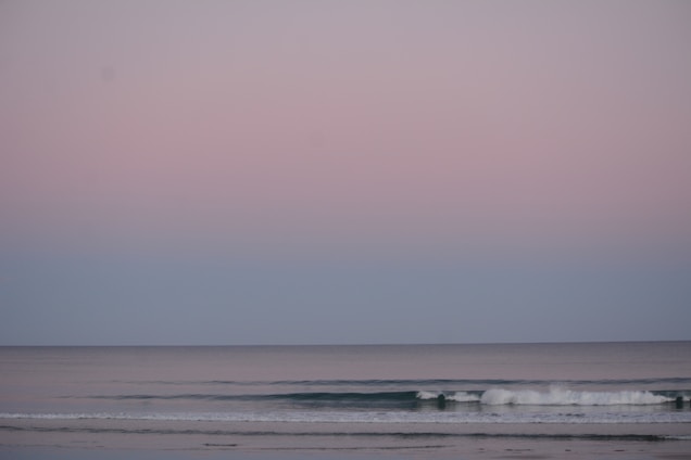 A serene seascape with calm ocean waves gently rolling towards the shore under a pastel-colored sky. The horizon divides the soft pink and blue hues of the sky and the muted tones of the sea.