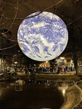 A large illuminated globe resembling Earth is suspended above a park setting at night. The globe is surrounded by leafless tree branches and is reflected in a nearby pond. A crowd of people gathers around the pond, illuminated by nearby streetlights, offering a mix of natural and artificial light sources.