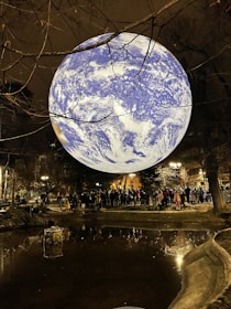 A large illuminated globe resembling Earth is suspended above a park setting at night. The globe is surrounded by leafless tree branches and is reflected in a nearby pond. A crowd of people gathers around the pond, illuminated by nearby streetlights, offering a mix of natural and artificial light sources.