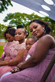 A group of women and a child are seated, wearing elegant pink dresses with lace and floral details. They appear to be attending an outdoor event, perhaps a wedding or celebration, as suggested by their formal attire. The background features green foliage, indicating a lush, natural setting, with a draped fabric providing shade.