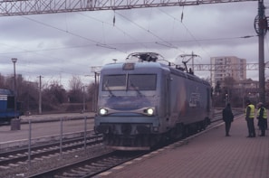 A train is stationed on a railway track surrounded by overhead electrical lines. Three people are standing on the platform, two of whom are wearing high-visibility vests. The scene is set against a cloudy sky and an urban backdrop with trees and apartment buildings in the distance.