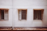 Three windows with wooden shutters are evenly spaced along a plain, light-colored wall. The shutters are partially open, revealing metal bars behind each window. The ground in front of the wall is slightly weathered.