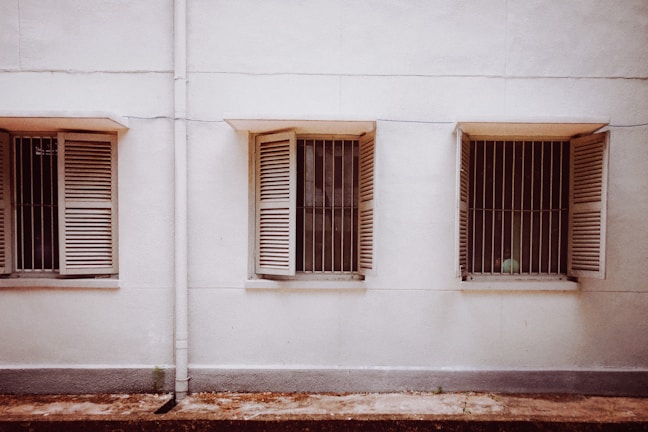 Three windows with wooden shutters are evenly spaced along a plain, light-colored wall. The shutters are partially open, revealing metal bars behind each window. The ground in front of the wall is slightly weathered.