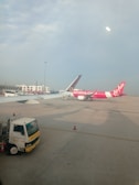 Two airplanes are parked on the tarmac of an airport. One airplane features a predominantly pink color with the AirAsia logo visible. The background includes airport buildings and infrastructure, with a clear blue sky overhead. A maintenance truck is positioned in the foreground beside the airplane.