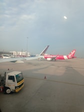 Two airplanes are parked on the tarmac of an airport. One airplane features a predominantly pink color with the AirAsia logo visible. The background includes airport buildings and infrastructure, with a clear blue sky overhead. A maintenance truck is positioned in the foreground beside the airplane.