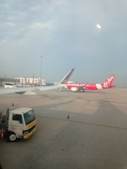 Two airplanes are parked on the tarmac of an airport. One airplane features a predominantly pink color with the AirAsia logo visible. The background includes airport buildings and infrastructure, with a clear blue sky overhead. A maintenance truck is positioned in the foreground beside the airplane.