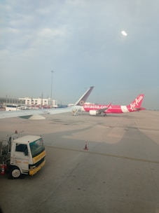 Two airplanes are parked on the tarmac of an airport. One airplane features a predominantly pink color with the AirAsia logo visible. The background includes airport buildings and infrastructure, with a clear blue sky overhead. A maintenance truck is positioned in the foreground beside the airplane.