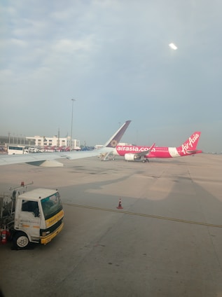 Two airplanes are parked on the tarmac of an airport. One airplane features a predominantly pink color with the AirAsia logo visible. The background includes airport buildings and infrastructure, with a clear blue sky overhead. A maintenance truck is positioned in the foreground beside the airplane.