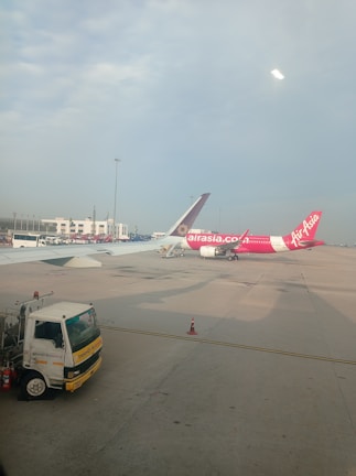 Two airplanes are parked on the tarmac of an airport. One airplane features a predominantly pink color with the AirAsia logo visible. The background includes airport buildings and infrastructure, with a clear blue sky overhead. A maintenance truck is positioned in the foreground beside the airplane.