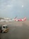 Two airplanes are parked on the tarmac of an airport. One airplane features a predominantly pink color with the AirAsia logo visible. The background includes airport buildings and infrastructure, with a clear blue sky overhead. A maintenance truck is positioned in the foreground beside the airplane.