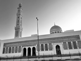 A large mosque with an intricately designed facade features a tall minaret and a prominent dome with decorative patterns. The architecture includes arches, Arabic script, and geometric designs. A metal fence lines the front, and a streetlight is visible in the foreground.