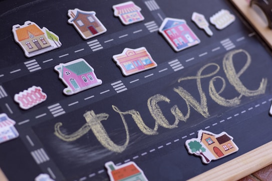A chalkboard with a road drawn on it, featuring small cut-out illustrations of various houses and shops placed along the road. The word 'travel' is written in yellow chalk in a large, cursive style, occupying the central part of the board.