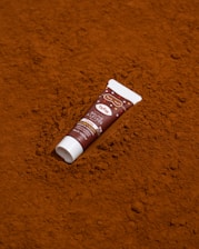 A tube of lip balm is placed on a textured surface that resembles cocoa powder. The design of the tube includes brown and white colors with the text 'TRÈSW' and 'Baume à lèvres' indicating it is a cocoa-flavored balm.