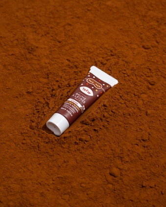A tube of lip balm is placed on a textured surface that resembles cocoa powder. The design of the tube includes brown and white colors with the text 'TRÈSW' and 'Baume à lèvres' indicating it is a cocoa-flavored balm.