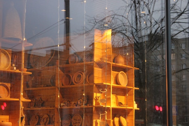 A wooden shelving unit contains a variety of handcrafted wooden items, possibly kitchenware or decorative pieces, displayed inside a store. The scene reflects an outdoor cityscape with trees and buildings visible through the glass, creating a blend of interior and exterior environments.