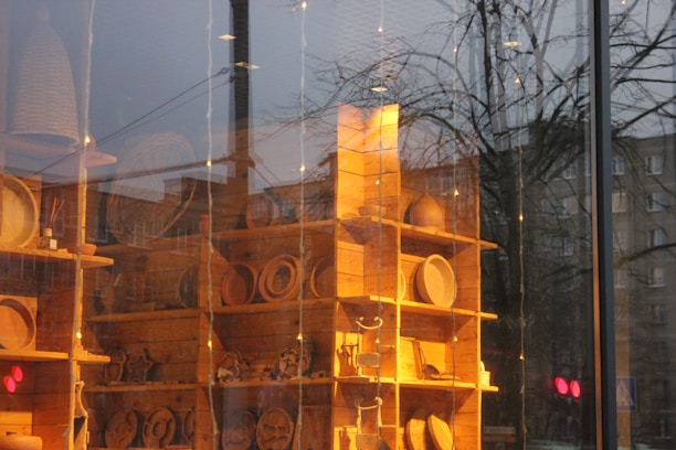A wooden shelving unit contains a variety of handcrafted wooden items, possibly kitchenware or decorative pieces, displayed inside a store. The scene reflects an outdoor cityscape with trees and buildings visible through the glass, creating a blend of interior and exterior environments.