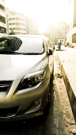 A silver car is parked on the side of a street bathed in bright sunlight. The focus is on the car's front and side, highlighting its clean lines and shiny finish. The background shows an urban setting with tall buildings and a slightly blurred street scene, creating a warm, glowy effect.