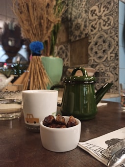 A cozy indoor setting featuring a table with a small white bowl of dates, a white mug with a car illustration, and a dark green teapot. The background includes a decorative wall with intricate patterns and a bundle of dried wheat placed in a green vase.