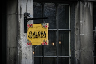 A vibrant yellow wooden sign displays the text 'Aloha Espresso Bar' with a pineapple graphic. The sign features tropical floral decorations at the corners and hangs on an iron bracket against a stone wall. Dimly lit windows behind the sign add to the ambiance.