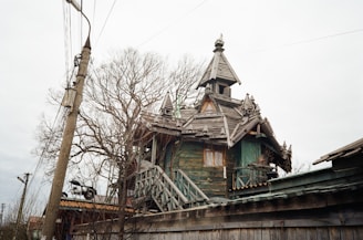 A rustic wooden house with intricate architectural details and a steep roof, surrounded by bare trees and power lines. The structure appears old and weathered, lending a historical or vintage feel.